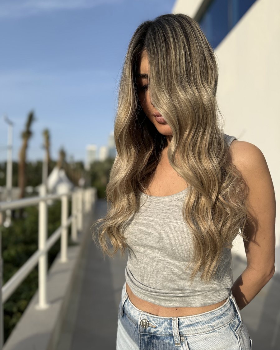 Long hair with a dark root and a beige-blonde balayage, styled in even waves past the shoulders.