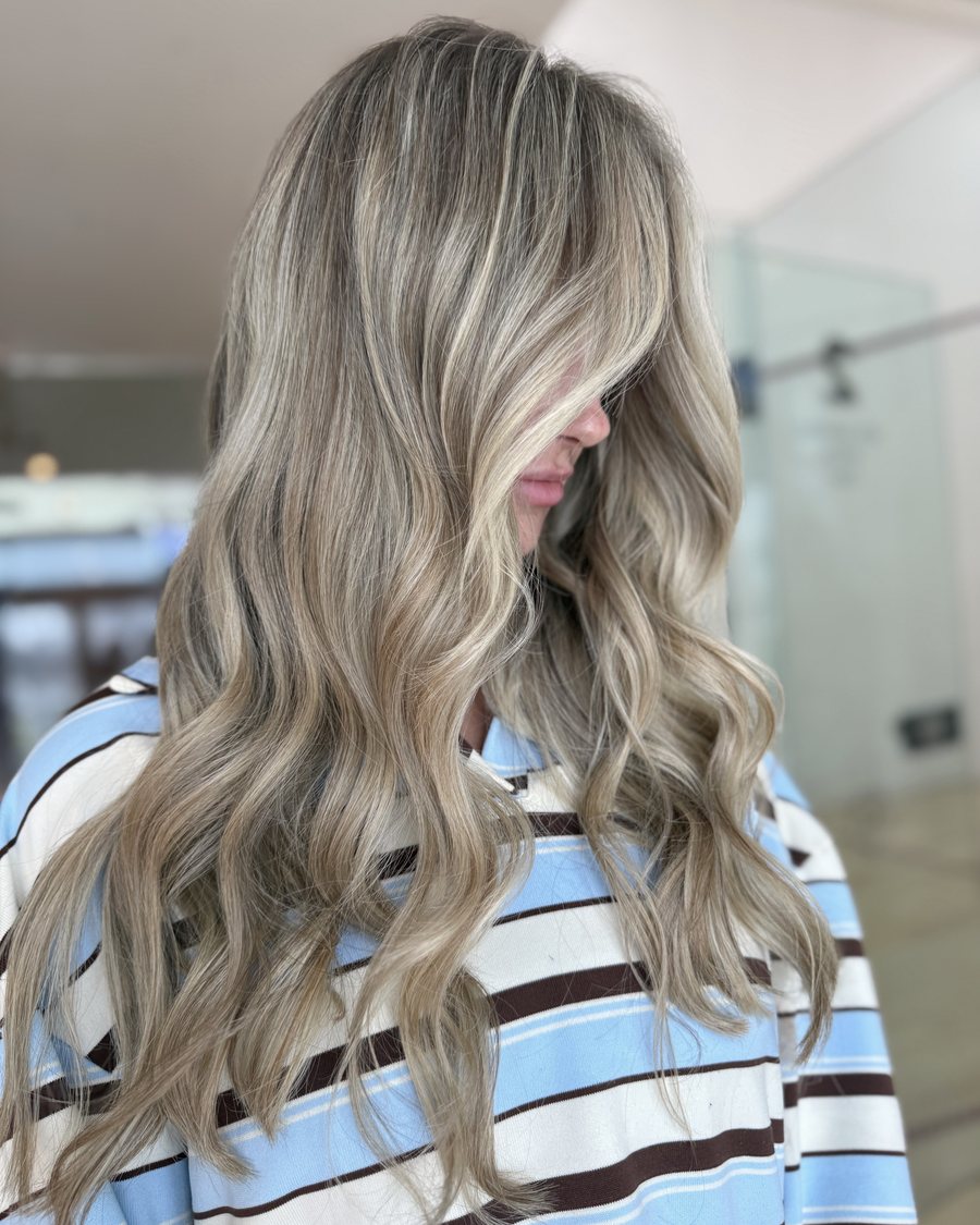 Long hair with cool ash-blonde highlights over a darker root, styled in soft waves and swept across one side.